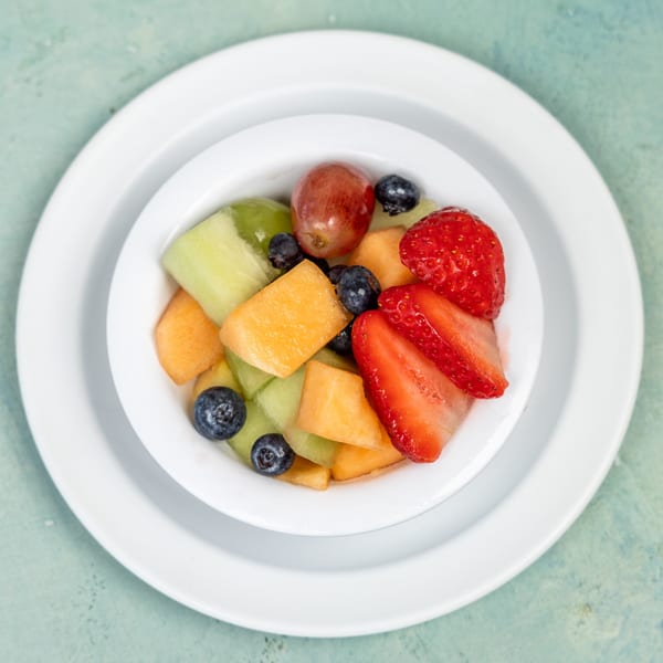 A white bowl filled with fresh fruit salad, including strawberries, blueberries, grapes, cantaloupe, and honeydew.