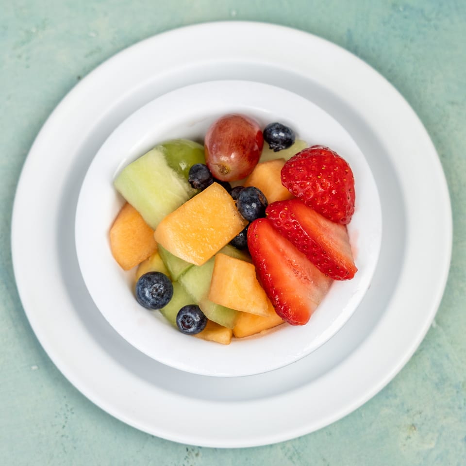 A white bowl filled with fresh fruit salad, including strawberries, blueberries, grapes, cantaloupe, and honeydew.