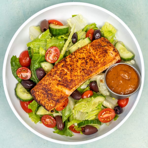 A grilled salmon fillet on a bed of fresh green salad with tomatoes, cucumbers, and olives.