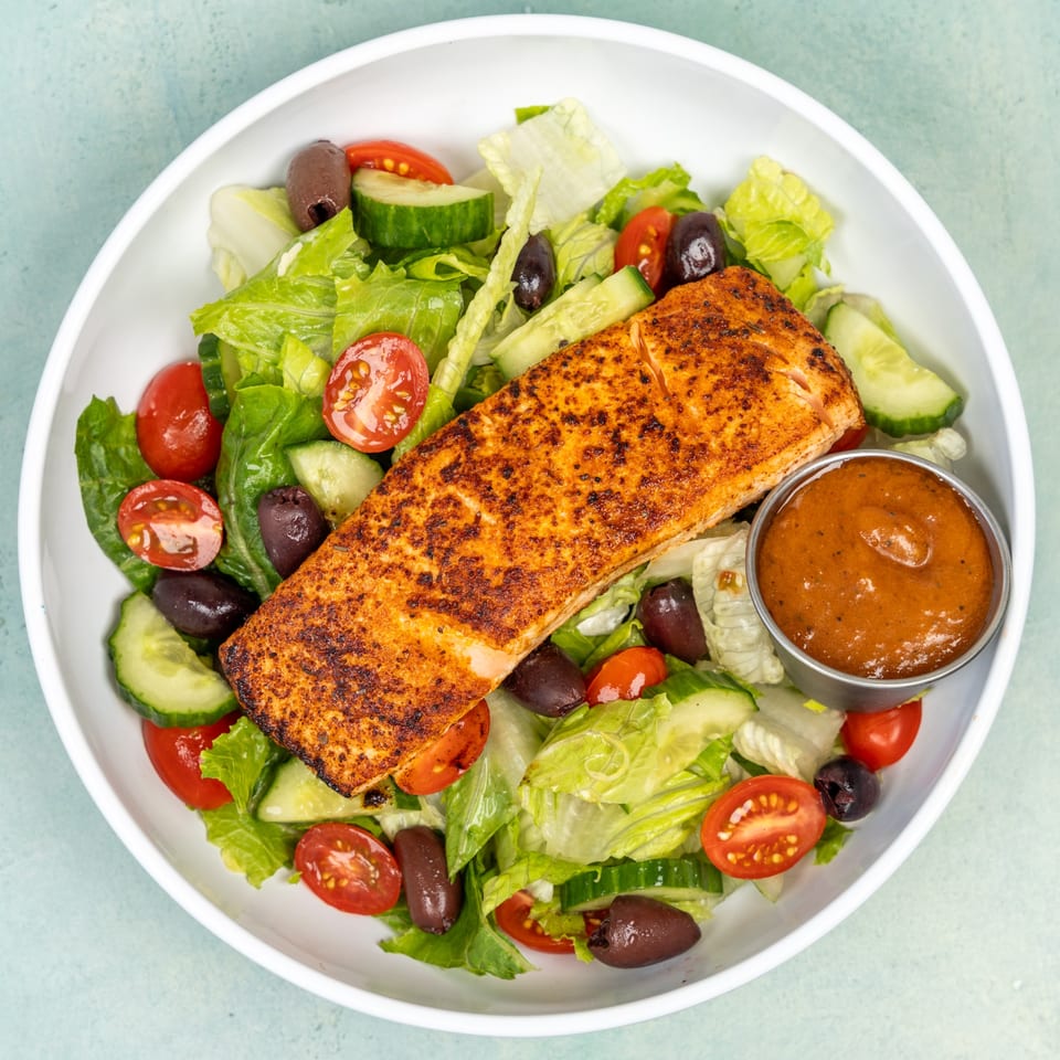 A grilled salmon fillet on a bed of fresh green salad with tomatoes, cucumbers, and olives.