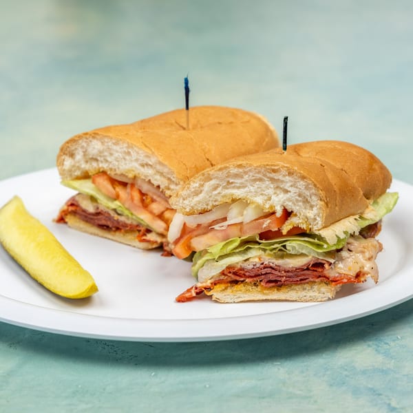 Two halves of a submarine sandwich with meat, cheese, lettuce, tomato, and onion, with a pickle spear.